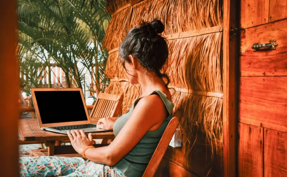 Back view of woman sitting in front of computer outside eco-friendly straw house building guesthouse.